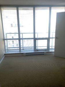 bright empty room with large floor-to-ceiling windows and white blinds allowing ample natural light, beige carpeting, and a simple white door, offering a clean and versatile living space with city views