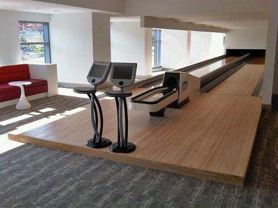 modern indoor bowling alley with sleek wooden lanes, digital scoring monitors, and contemporary seating featuring a bright red sofa and white side table, illuminated by ample natural light from large windows, creating a clean and inviting recreational space.