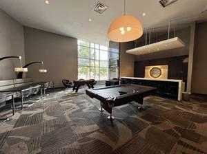 modern game room featuring a stylish pool table, large windows allowing natural light, cozy seating area with black leather couches, and contemporary lighting fixtures, creating a sleek, inviting entertainment space with a sophisticated ambiance.