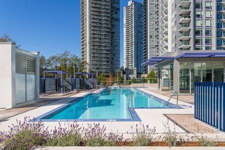 modern outdoor swimming pool area surrounded by high-rise residential buildings under a clear blue sky, featuring clean pool decks, purple pergolas, and landscaped lavender plants, offering a serene and upscale urban living environment